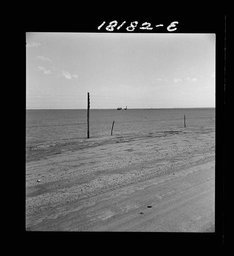 The image is in black and white, depicting a desolate landscape with an expansive body of water on the horizon under a vast sky. There are multiple poles sticking out from the ground at varying angles, indicating some form of fencing or barrier that has been eroded by weather conditions over time. The sandy terrain shows signs of wear, suggesting prolonged exposure to elements like wind and possibly saltwater from proximity to the sea. In the distance, there is a faint outline of what appears to be another structure or vessel on the water surface. There's no human activity visible in this scene; it conveys an atmosphere of isolation and abandonment. The photograph has alphanumeric text at the top left corner reading "18/32-6", which might refer to a cataloging system used by the photographer or archive that holds the image.