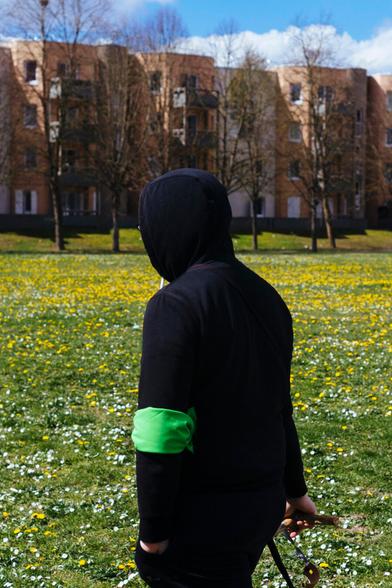 La photographie d'un jeune homme encapuchonné marchant dans un pré fleuri. A l'arrière plan des barres d'immeubles.