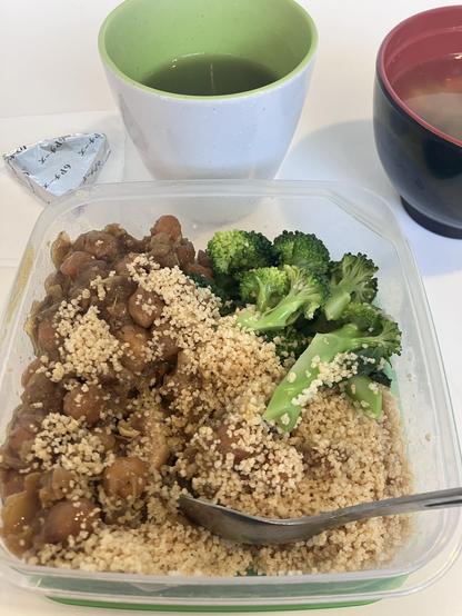A meal in a transparent container featuring couscous topped with chickpeas and a side of steamed broccoli. Accompanying the meal are a green beverage in a white mug and a black bowl with a lighter colored liquid. A wrapped portion of processed cheese is also visible