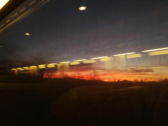 Photographie pris depuis l'intérieur d'un train, par la fenêtre.
Le paysage se compose d'un pré simple, avec quelques arbres en fond. Le ciel est rose-orangé, incendiaire.
On voit en surimpression les lumières du train qui se reflètent dans la vitre, ça fait un rang de lumière jaune et carre par-dessus le ciel (qui est un peu sale)