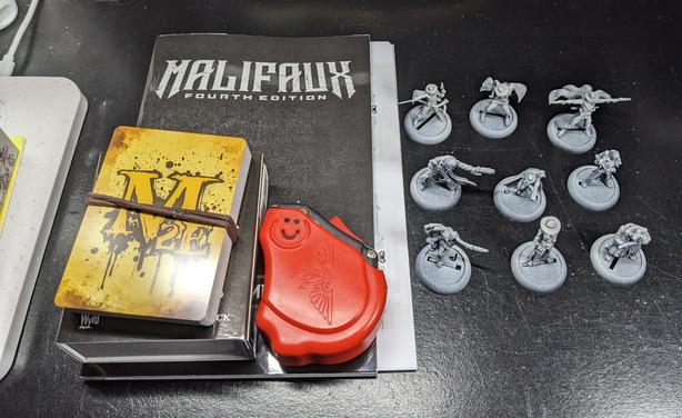 The same models as the previous photo sitting next to a rulebook for Malifaux fourth editions, a tape measure, a deck of gate cards and a box of tokens.