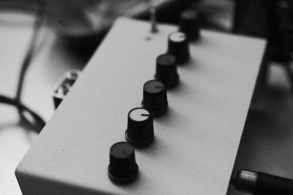 A home-made electronic device with six potentiometer knobs in a row across the top. The power switch is there, too, but it is blurry. Background is a cluttered table top, all pretty blurry.
