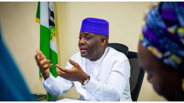 Tunji Alausa sits at a desk wearing a white outfit and a purple cap, gesturing with both hands during a discussion. A Nigerian flag is visible behind him, and another person is partially visible in the foreground.