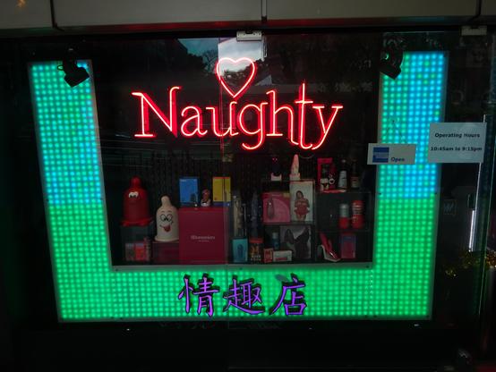 Ein Schaufenster mit einer hellgrün leuchtenden Umrahmung aus kleinen quadratischen Lichtern. In der Mitte des Schaufensters steht in roter Neonbeleuchtung das Wort 'Naughty' mit einem Herzsymbol über dem Buchstaben 'u'. Darunter sind verschiedene Produkte in Regalen angeordnet, darunter Flaschen und Verpackungen in unterschiedlichen Farben. Am unteren Rand des Schaufensters leuchten chinesische Schriftzeichen in Blau und Lila. Rechts neben dem Schaufenster ist ein Schild mit den Öffnungszeiten von 10:45 Uhr bis 21:15 Uhr zu sehen, daneben ein kleines Schild mit der Aufschrift 'Open'.