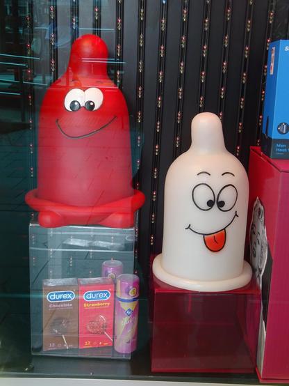 Zwei große, bunte Figuren in Form von Kondomen mit Gesichtern stehen nebeneinander. Die linke Figur ist rot mit großen, weißen Augen und einem breiten Lächeln. Die rechte Figur ist weiß mit großen, schwarzen Augen, die nach unten schauen, und einer herausgestreckten roten Zunge. Unter der roten Figur befinden sich zwei Packungen Durex-Kondome in den Sorten Chocolate und Strawberry sowie eine pinkfarbene Verpackung mit einem Kondom darauf.
