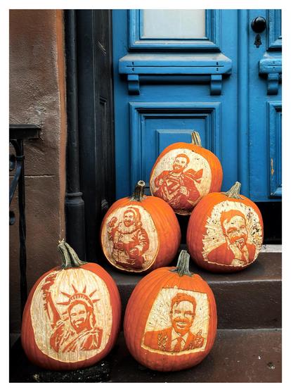 Photo of five large carved pumpkins on the steps in front of a blue, paneled exterior door in a brownstone building. The carvings are all images of  a young, smiling bearded man (NYC’s new Mayor-Elect Zohran Mamdani) in various whimsical scenarios: as the face of the Statue of Liberty; as Santa Claus; ripping open his shirt Superman-style to reveal a big “Z” underneath.