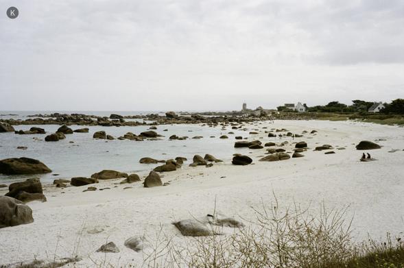 Strand mit hellem Sand und verstreuten Felsen an einer ruhigen, flachen Bucht; bewölkter Himmel, wenige Häuser und ein Sémaphore in der Ferne, zwei Personen sitzen am Ufer.