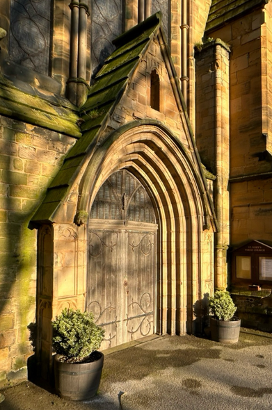A church door with decorative surrounding architecture, dappled with shadows cast by golden sunlight an hour before sunset.
