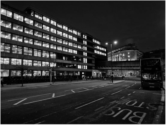 Night time photo of a large office building on the other side of the street with a London two decker bus on the right.  The driver of the bus is lit up.