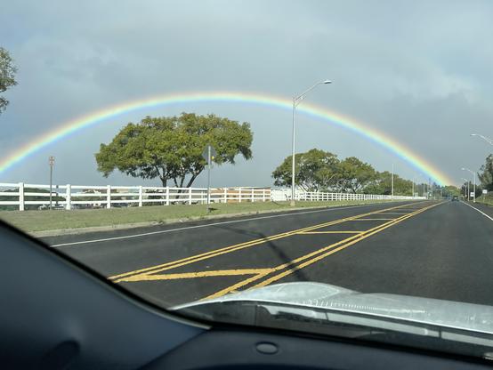 First of four photos taken from a moving vehicle of a large rainbow stretching across the photo frame.  Some scattered wide-canopied trees and a white picket fence lining the roadway are enclosed underneath the rainbow.