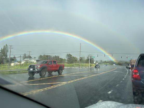 Second of four photos taken from a moving vehicle of a large rainbow stretching mostly across the photo frame.  A very faint double rainbow is visible in the gray sky above the main rainbow.  The road pavement is wet from the rain and a large red pickup truck is framed underneath the rainbow.