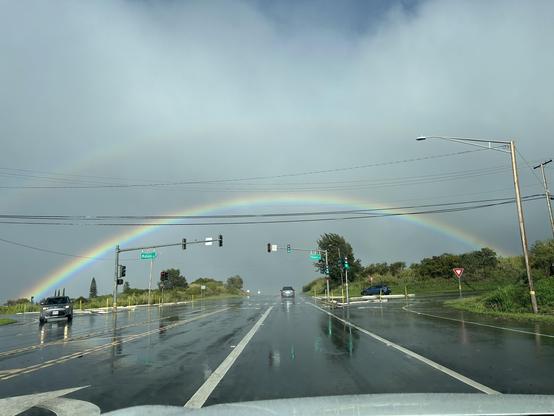 Third of four photos taken from a moving vehicle of a large rainbow stretching across the photo frame but seemingly further in the distance than the previous photos.  The wet highway stretches straight ahead, disappearing into the horizon directly beneath the rainbow.  Horizontal powerlines cross the highway in front of the rainbow, creating a geometric effect of the straight powerlines intersecting the curved rainbow.