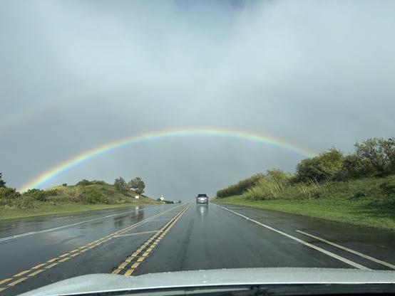 Fourth of four photos taken from a moving vehicle of a large rainbow stretching across the photo frame.  As in the third photo the highway stretches toward the horizon under the rainbow, and a lone car in the distance appears about to cross directly under the vibrantly colored bow.