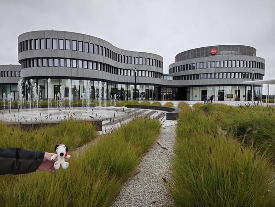 Häkelsnoopy besichtigt die Leica Welt in Wetzlar, hinter ihm das Ausstellungs und Produktionsgebäude, dessen Form einem Objektiv nachempfunden ist. Die Brunnenanlage davor imitiert die Lamellen eines Kameraverschlusses.