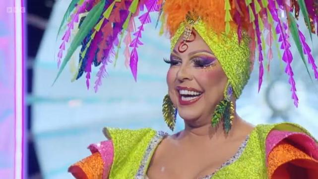 La Voix, British drag queen superstar in a screenshot from RuPaul's Drag Race UK. It's a close-up of her face, and she's wearing a large Carmen Miranda-style headpiece with many feathers and feathery earrings.
