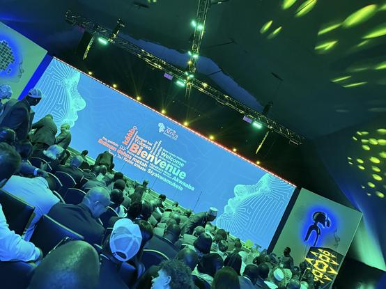 A large audience sits facing a brightly lit stage with a blue screen displaying multilingual welcome messages at a technology conference. The atmosphere is vibrant, with green and blue lighting.