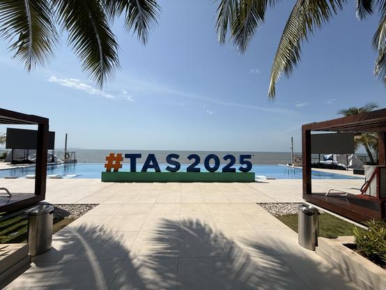 A large “#TAS2025” sign stands by an infinity pool overlooking the ocean, framed by palm trees and two wooden cabanas under a clear blue sky.