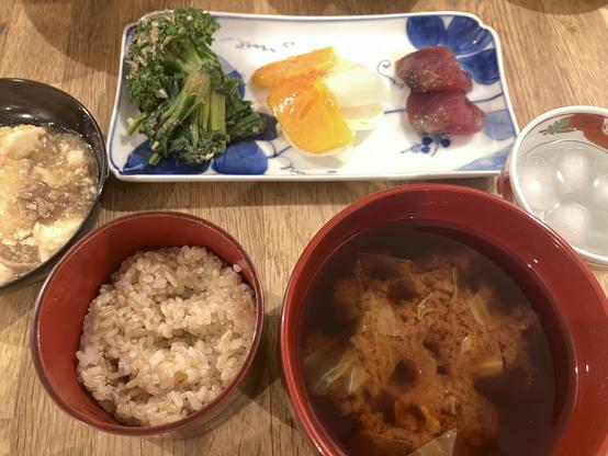 A Japanese meal featuring a bowl of miso soup, a portion of brown rice, sautéed greens, fresh slices of fish, and a carpaccio of Kaki and turnip, accompanied by a glass of awamori and water.