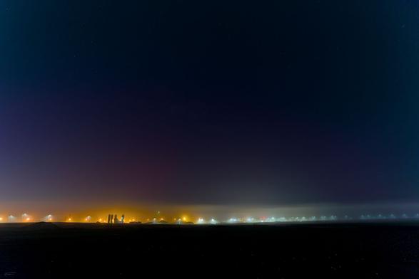 In dieser Nachtaufnahme verschmilzt die Stille der Dunkelheit mit einem Hauch von geheimnisvoller Aktivität. Der Himmel, tief und fast schwarz, wird am linken Horizont von einem zarten, violett schimmernden Schein umgeben, ein schwacher Abglanz von Polarlichtern in dieser Nacht.
Am Horizont zieht sich eine dünne, gelblich-orangefarbene Schicht hin, die von den Lichtern einer fernen Siedlung oder Baustelle stammt. Diese künstlichen Lichtquellen tauchen den unteren Teil des Bildes in einen leichten Dunst, als würde der Nebel die Lichter sanft verschlucken. Die Landschaft darunter wirkt flach und weitläufig, fast wie eine endlose Ebene, die sich in die Dunkelheit erstreckt.
Die Lichter – mal punktuell, mal flächig – erzählen von menschlicher Präsenz in dieser ansonsten stillen Nacht. Sie schaffen einen Kontrast zwischen der natürlichen Weite des Himmels und der bescheidenen, aber spürbaren Aktivität am Boden.