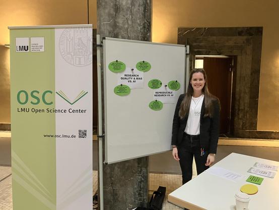 An image of a female presenter wearing a white jersey under a black blazer and black pants stands in front of a whiteboard with white and green papers stuck on to it. To the left of the board is a green and white banner with the words “OSC; Open Science Center”. To the right of the speaker, a table can be seen with papers and a coffee cup on it.