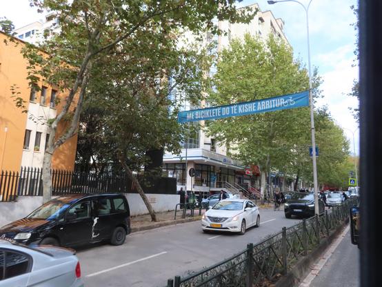 View from a public bus window across the traffic median to a light blue banner with white text and the icon of an upright bicycle. It says "Me biçikletë do të kishe arritur". Traffic is light because it's about 11am.