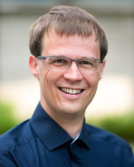 Mann mit Brille und dunkelblauem Hemd lächelt in die Kamera, der Hintergrund ist unscharf. Foto: Lucas Adrian/erweitert durch KI