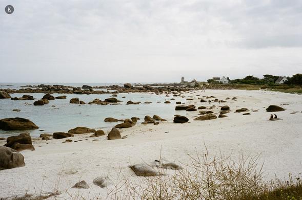 Strand mit hellem Sand und verstreuten Felsen an einer ruhigen, flachen Bucht; bewölkter Himmel, wenige Häuser und ein Sémaphore in der Ferne, zwei Personen sitzen am Ufer.