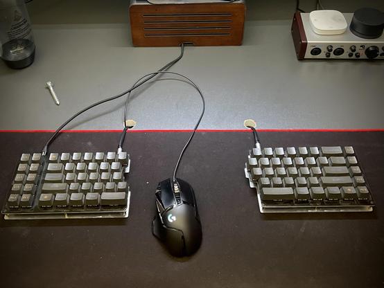 Ein Foto eines Arbeitstisches. In der unteren Hälfte eine geteilte Tastatur, zwischen den beiden Hälfte  eine Computermaus.