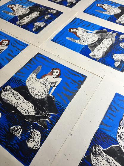 Several of my 11” x 14” selkie linocuts on cream coloured Japanese washi paper with speckled bark inclusions are laid out on the table to dry. Each print shows a red-headed selkie shedding her seal skin in a rock in the dark blue sea surrounded by grey spotted harbour seals. The light blue sky behind has grey clouds.