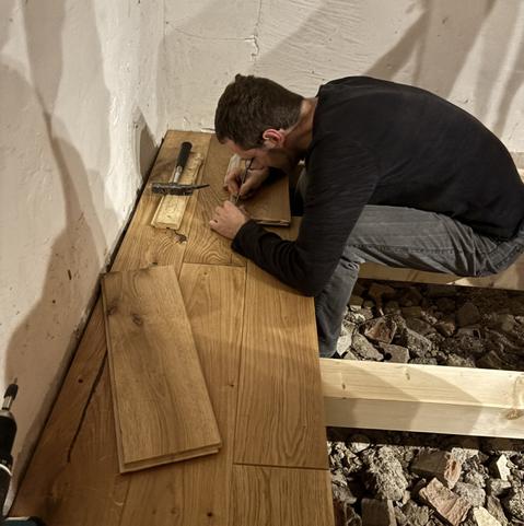 Die ersten drei Dielenreihen liegen schon, darauf liegt weiters loses Material und Werkzeug. Ein Mann sitzt auf einen Balken und zeichnet Schnittkanten an.