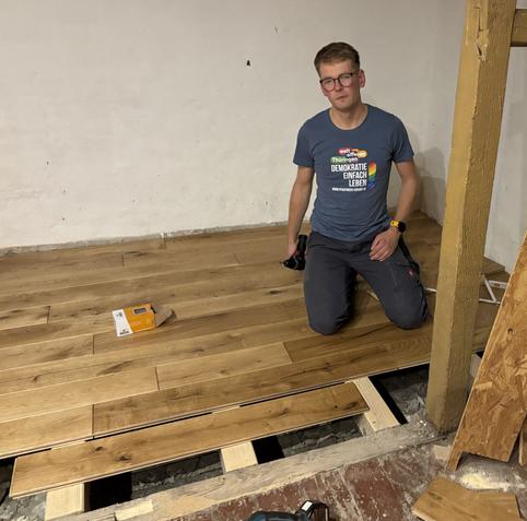 Ein Mann sitzt auf dem schon verlegten Dielen und vor ihm liegt eine lose die er befestigen möchte um die letzten zwei Reihen zu schließen.