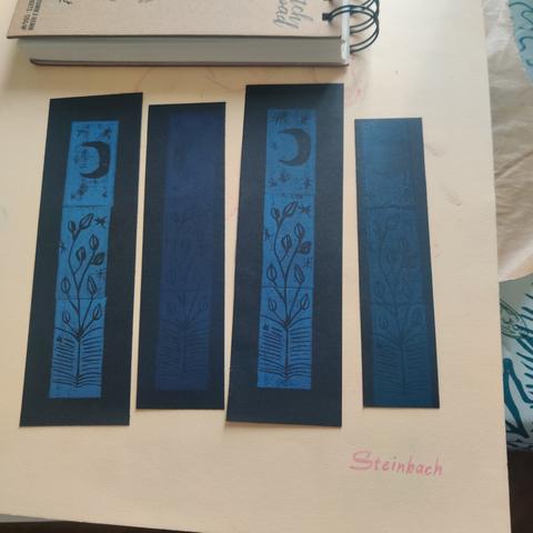 Black paper bookmarks with various shades of blue ink prints depicting a moon, stars, a climbing plant and a book.