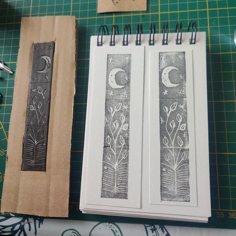 The same print (moon, stars, plant, book) but on white paper with black ink. Next to it on the left you can see the cut erasers and a makeshift cardboard shape for alignment.