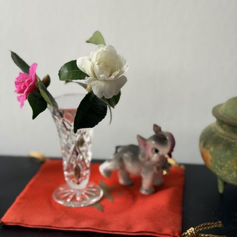 A small crystal bud vase holds two small camellia flowers, both with a few dark green leaves attached. One is white and the other pink. The vase sits on a red and gold cloth mat with gold tassles, with a cute elephant figurine and copper/gold incense burner nearby.