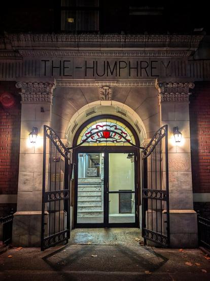 Night-time view of the entrance to The Humphrey building. The entrance features an arched doorway with ornate stonework and a stained-glass window above the door. The building's name, 'THE HUMPHREY,' is prominently displayed in capital letters at the top of the arch. The entrance is framed by two black wrought-iron gates, which are open, revealing a glass door and a staircase inside. The scene is illuminated by two wall-mounted lanterns on either side of the entrance, casting a warm glow on the stone and brick façade.