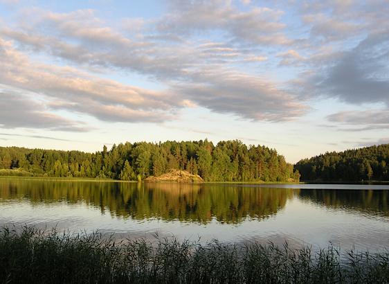 A lake view with forest on the other bank lit by the evening sun and reflected in the calm waters. The sky is partially covered with light clouds, also highlighted by the sun.