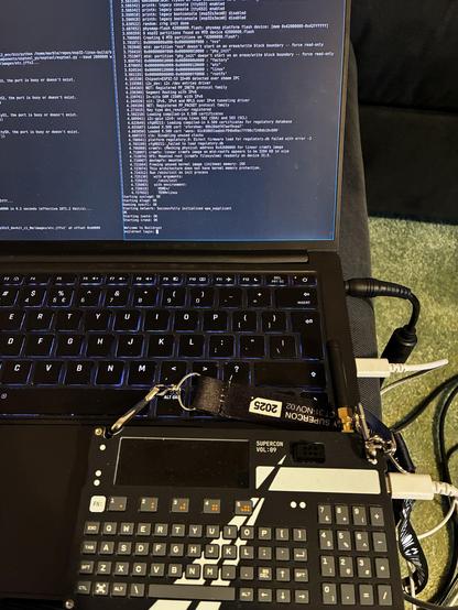 A laptop with a visible keyboard, displaying terminal commands and code on the screen. Attached below is a device labeled "SUPERCON VOL. 89," featuring a keyboard and antennas, connected with cables to the laptop.