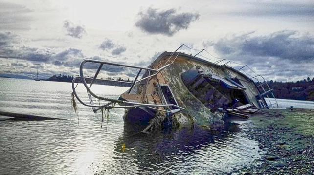 A weathered boat is partially submerged in water, tilted on its side near a rocky shore under a cloudy sky. The boat's metal frame and side are heavily corroded. Parts of its interior are visible, including stairs. Railings extend around the boat's edges. In the background, across the water, are trees and a distant bridge. A small sailboat is also visible in the distance.