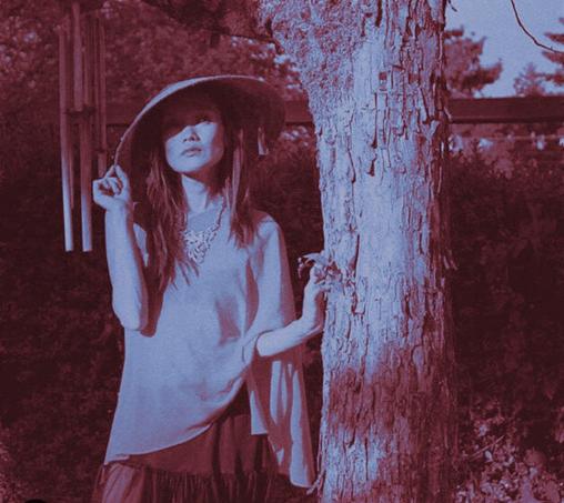 A woman with long, auburn hair, wearing a wide-brimmed hat, a light-colored blouse, and a tiered skirt, stands next to a large tree. She looks upward with a contemplative expression, and her right hand gently touches the bark. A wind chime hangs in the background to the left. The image has a duotone effect, using red and blue hues.