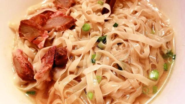 Close-up shot of a bowl of soup noodles with char siu pork and scallions; the wide noodles are pale, the pork has a red hue, and the soup is a light brown color. The bowl is white. The shot is well lit.