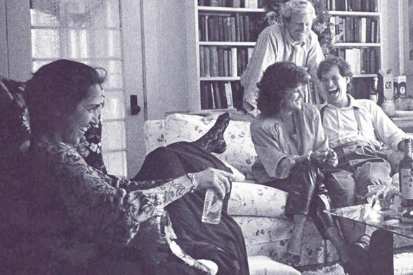 1987 Vintage Indoor Gathering in a Living Room

The image captures an indoor scene with four individuals engaged in what appears to be a jovial conversation.
One person is seated on the left, seemingly relaxed and possibly smoking or holding something near their mouth.
In the middle ground, two more people are present; one sits beside another standing individual who seems older, all three appear content and involved in discussion.
The background features bookshelves filled with books, suggesting a homey environment that could be indicative of intellectual pursuits.
