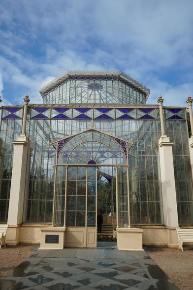 A large, ornate glass structure with a light beige frame stands against a blue sky with scattered white clouds. The building is predominantly glass, with a complex network of panes and beams creating a Victorian-era greenhouse appearance. The structure has a central arched entryway flanked by symmetrical rows of vertical glass panels and supporting columns. A small, dark-colored plaque is visible above the entryway. The ground in front of the building is paved with dark gray stone tiles, and glimpses of foliage can be seen through the glass.

Provided by @altbot, generated privately and locally using Gemma3:27b