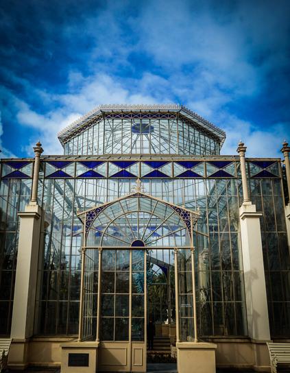 A large, ornate glass structure with a light beige frame stands against a blue sky with scattered white clouds. The building is predominantly glass, with a complex network of panes and beams creating a Victorian-era greenhouse appearance. The structure has a central arched entryway flanked by symmetrical rows of vertical glass panels and supporting columns. A small, dark-colored plaque is visible above the entryway. The ground in front of the building is paved with dark gray stone tiles, and glimpses of foliage can be seen through the glass. Provided by @altbot, generated privately and locally using Gemma3:27b