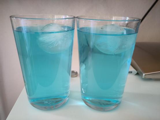 Auf dem Bild stehen zwei identische, durchsichtige Trinkgläser nebeneinander auf einer hellen Oberfläche. Beide Gläser sind bis fast zum Rand mit einem leuchtend türkisblauen Getränk gefüllt. In jedem Glas schwimmt eine große, runde Eiskugel nahe der Oberfläche. Im Hintergrund steht ein Laptop auf der rechten Seite. Die Szene wirkt ruhig und ordentlich, und die kräftige blaue Farbe der Getränke fällt besonders ins Auge.