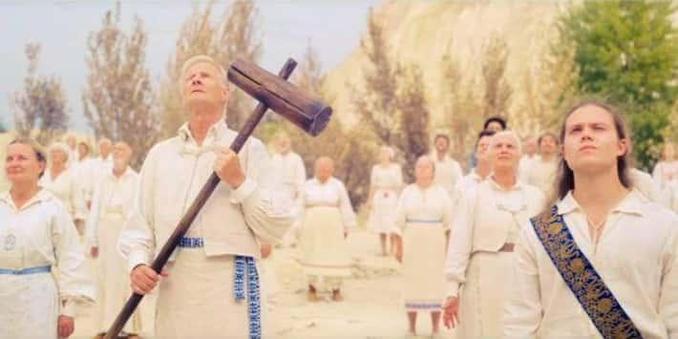 Eine Gruppe Menschen in weißen Gewänden schaut nach oben. Einer trägt einen sehr überdimensionierten Holzhammer mit einem langen Stiel mit beiden Händen