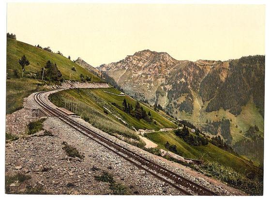 The image depicts a picturesque landscape featuring the Rochers de Naye mountain range, specifically from an old postcard or print. It shows railroad tracks winding along a steep hillside with lush greenery and scattered trees. The background showcases rugged mountainside terrain covered in grass and shrubs, with patches of rocky outcrops visible at higher elevations. A small path can be seen curving through the landscape parallel to the train tracks on lower slopes.

The photograph's vintage quality is evident from its yellowish tone and fine grain texture characteristic of early colorized images produced using a process like photochrom or similar techniques prevalent in that era (circa 1890-1900). The image provides insight into transportation infrastructure during the late 19th century, particularly highlighting railway developments connecting mountainous regions.

The overall atmosphere conveyed by this scenery is one of serene natural beauty juxtaposed with human engineering. It captures a moment in Swiss history when rail travel began to transform remote areas like those around Lake Geneva (Lac Léman) into accessible destinations for travelers and tourists alike.