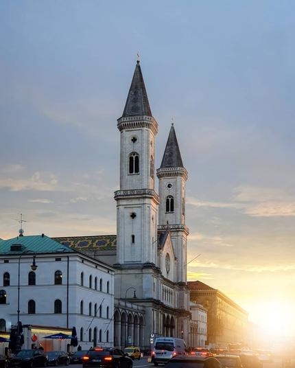 A twin-towered church building in the city at sunset, with warm light shining down the street and cars driving in the foreground.