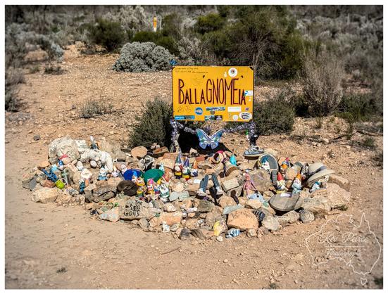 A wide angle photo of a yellow wooden sign in a dry, scrubby roadside environment. The sign, hanging between two posts wrapped in fairy lights, reads "BALLAGNOMEIA" in large orange, whimsical text.
Below the sign is a large cairn or pile of rocks covered in dozens of small garden gnomes and various trinkets, including a blue butterfly decoration, a small toy goat, and some hats. The ground is dry dirt and gravel.