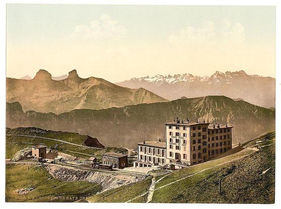 The image shows a picturesque landscape featuring an elevated viewpoint overlooking mountains with prominent peaks in the distance, some of which are partially covered by snow. In the foreground and middle ground, there is a large building labeled "Rochers de Naye Grand Hotel," indicating it's a hotel located on or near Rochers de Naye mountain range. Adjacent to this main structure, smaller buildings can be seen, possibly associated with the hotel such as service facilities or additional accommodations.

The terrain slopes gently upward from left to right in the foreground and middle ground where the hotel is situated, leading up into more dramatic mountainous regions further back. The sky above has a soft haze, suggesting either early morning light or atmospheric conditions typical of higher altitudes. The overall color palette consists primarily of muted earth tones with hints of greenery on the slopes.

In addition to the buildings and landscape, there are elements indicative of transportation: tracks leading away from the hotel could suggest railway lines, possibly linking this remote location to larger networks for travel purposes during its time period. This image seems to capture a moment in history where mountain resorts were becoming popular tourist destinations in Switzerland.