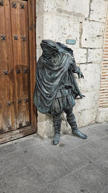 Escultura de bronce junto a la puerta de madera de la Casa de Zorrilla, en Valladolid. Representa a un caballero del Siglo de Oro, con capa, sombrero y botas altas, apoyado en la pared de piedra frente a la entrada del edificio histórico.